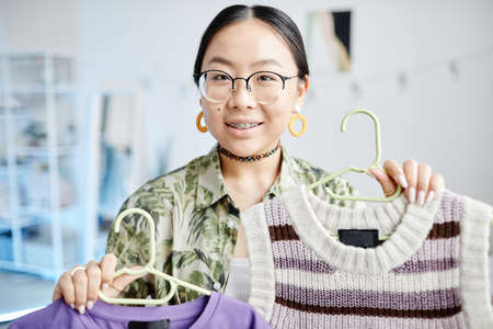 Portrait of Asian teenage girl with braces trying on different tops for trendy outfit and smiling at...