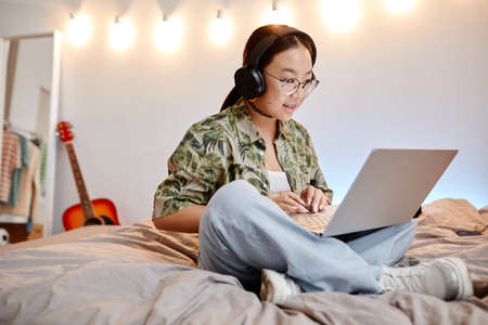 Full length portrait of Asian teenage girl using laptop while sitting on bed cross legged, copy...