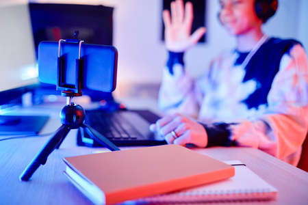 Closeup of teenage boy playing video game and streaming live in blue neon light waving at smartphone...