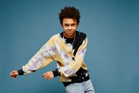 Minimal portrait of black teenage boy wearing tie dye shirt and dancing over blue background, copy...