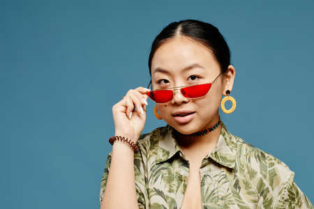 Minimal portrait of Asian teenage girl wearing trendy colored sunglasses over blue background, copy...