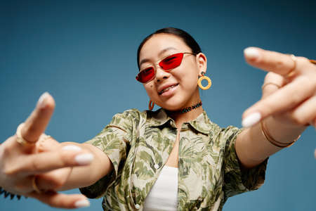 Portrait of trendy teenage girl wearing colored sunglasses and gesturing over vibrant blue backgroundの写真素材