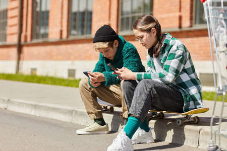 Couple of teens sitting n their skateboards outdoors and chatting online using smartphonesの写真素材