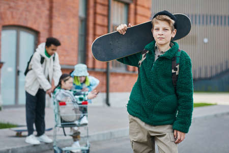 Portrait of skater boy holding his skateboard and looking at camera while standing on street with friends in backgroundの写真素材