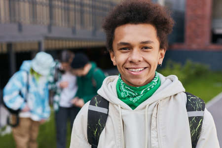 Portrait of African teenage boy with backpack behind his back smiling at camera standing outside of school buildingの写真素材