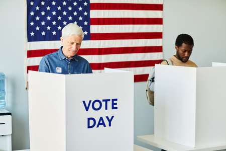 Portrait of two people in voting booths on election day against USA flag, copy space