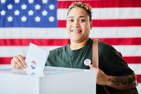 Portrait of smiling African American woman putting ballot in bin against USA banner background, copy...