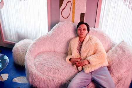 Portrait of Asian young man dressed in fur coat and party outfit relaxing in clu lounge lit by pink neon lightsの写真素材