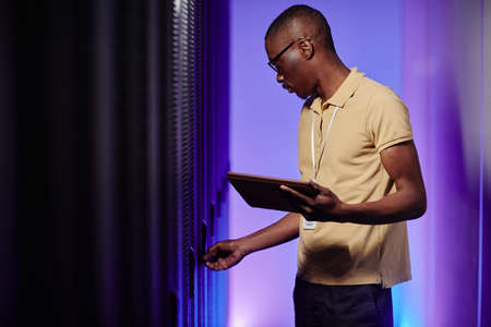 Side view portrait of system administrator inspecting data network in server room lit by neon light,...