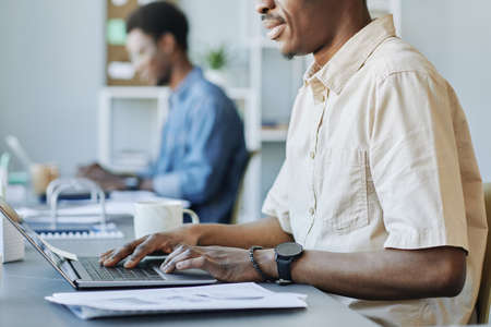 Cropped side view shot of adult black man typing at laptop while working in office and holding cup...