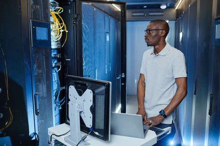 Side view portrait of black men setting up internet network and using laptop in server room lit by...