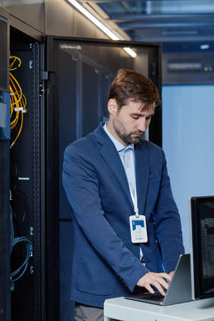 Portrait of Caucasian young man wearing blazer setting up internet network in server room and using laptopの写真素材