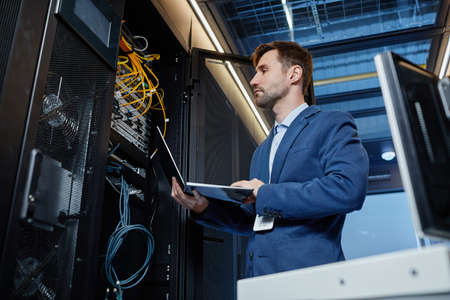 Low angle portrait of IT engineer holding laptop while setting up internet network in server roomの写真素材