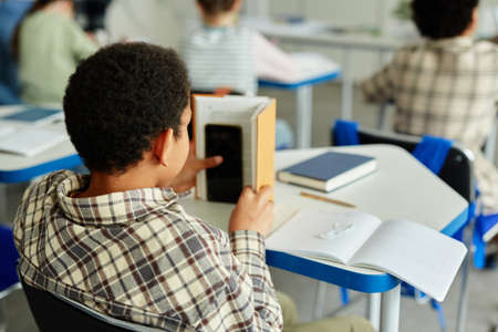 Back view at young African American kid hiding smartphone in book and cheating in classの写真素材