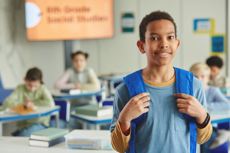 Waist up portrait of black young schoolboy with backpack looking at camera while standing in classroom, copy spaceの写真素材