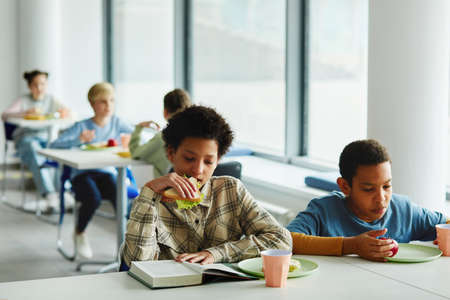 Portrait of schoolchildren at lunch break, focus on young african American girl eating sandwich and reading book, copy spaceの写真素材