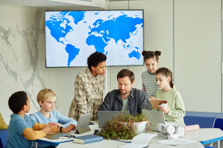 Diverse group of children with male teacher in modern school class on geography with map in...