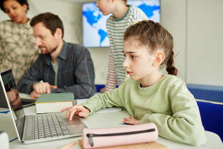 Side view portrait of cute schoolgirl using laptop in modern school classroom with kids in backgroundの写真素材