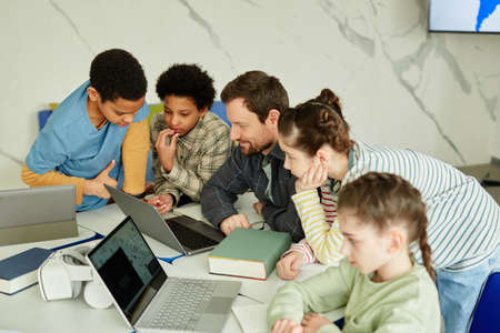 High angle portrait of smiling male teacher with diverse group of children using laptop together in...