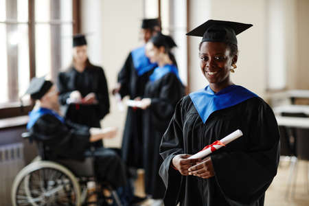 Waist up portrait of young African American woman wearing graduation gown and smiling at camera in classic universityの写真素材