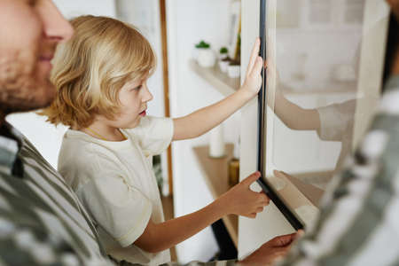 Side view portrait of cute blonde boy helping father hanging picture on wall while decorating new houseの写真素材