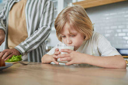Portrait of cute blonde boy drinking milk in morning with father cooking in cozy kitchen interior, copy spaceの写真素材