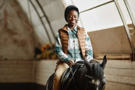 Portrait of smiling African American woman riding horse in indoor arena at horse ranch or practice stadium, copy spaceの写真素材