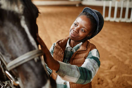 High angle portrait of young black woman caressing horse after sports practice in indoor riding arena, copy spaceの写真素材