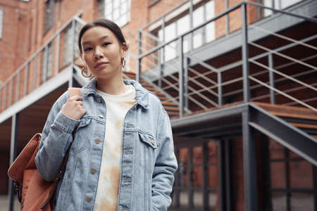 Pretty teenage girl in denim jacket holding backpack on shoulder while standing in front of camera against staircase and brick buildingの写真素材