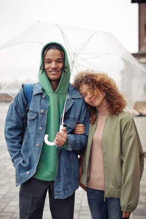 Happy young interracial couple in casualwear standing close to one another under umbrella and looking at camera during walkの写真素材