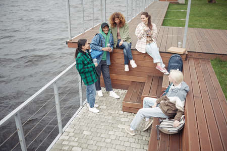 Group of multicultural teenage friends in casualwear having chat and relaxing by riverside while some of them sitting on wooden benchの写真素材