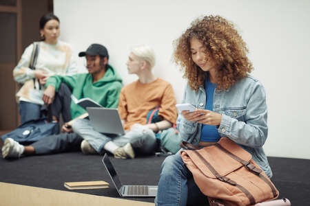 Pretty teenage girl in casualwear texting in smartphone at break while sitting in lecture hall...