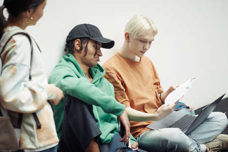 Two intercultural teenage guys looking through lecture notes in copybook held by one of them over laptop keypad while preparing for lessonの写真素材
