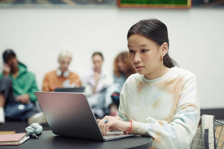 Asian teenage girl in white sweatshirt looking at laptop screen and touching keypad while working over school assignmentの写真素材