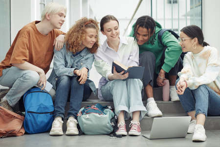 Large group of teenagers in casualwear reading open book held by smiling girl and discussing paragraph before lessonの写真素材