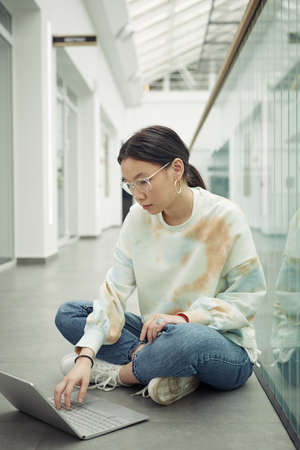 Serious teenager concentrating on network while sitting on the floor of corridor and looking for online information for new school projectの写真素材