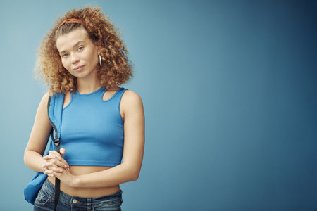 Pretty teenage girl with long thick wavy hair wearing blue crop top and jeans holding backpack on shoulder while standing by wallの写真素材