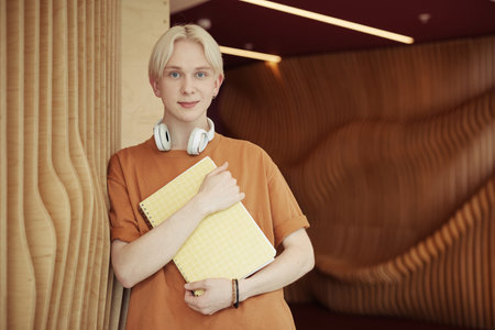 Smiling teeange guy with blond hair holding copybook by his chest while standing in front of camera...