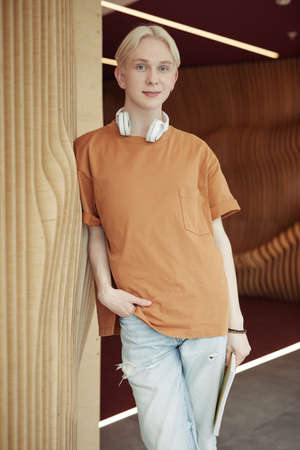 Smiling blond teenager in brown t-shirt and light blue jeans standing against wooden beige wall in...