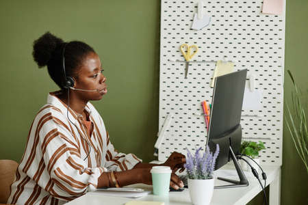 African young businesswoman in headphones sitting at her workplace with computer monitor and talking online with colleaguesの写真素材
