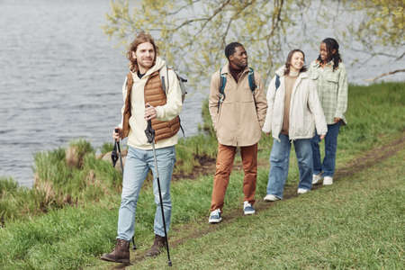 Young man walking with stick along the bank of the river together with his friends behind him