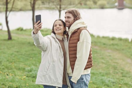 Happy young couple embracing and smiling at camera while making selfie portrait on mobile phone standing outdoorsの写真素材