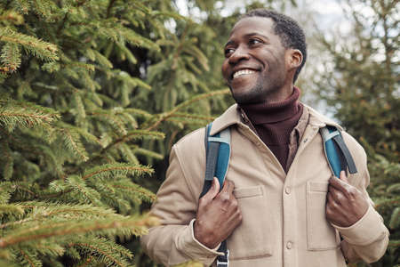 Happy African man with backpack enjoying his walk among fir trees in the park