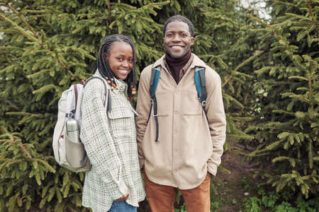 Portrait of african couple of hikers with backpacks smiling at camera while standing outdoors among fir treesの写真素材