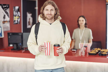 Portrait of young man with soda and popcorn looking at camera standing at cinema with snack bar in...