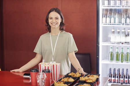 Portrait of young seller smiling at camera standing at counter and selling popcorn and nachos at cinemaの写真素材