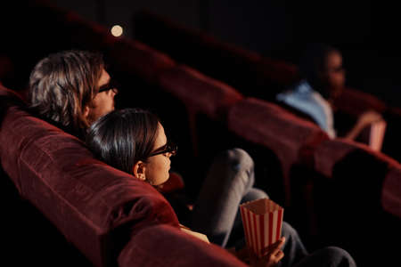 Rear view of young couple watching 3d movie in glasses on comfortable armchairs in cinemaの写真素材