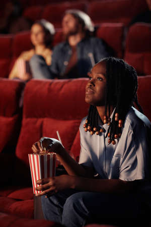 African young girl eating popcorn and watching movie on armchair in cinemaの写真素材