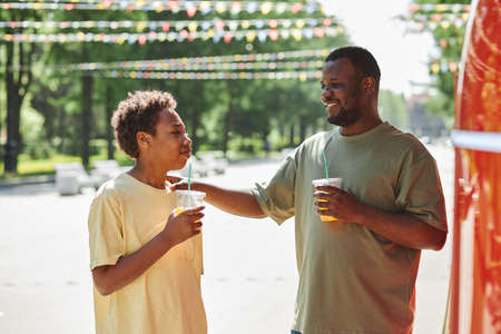 African smiling father drinking juice together with son during their stroll in the park in sunny dayの写真素材