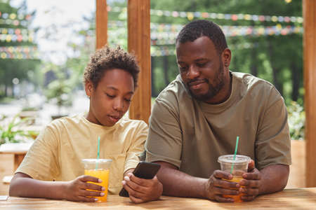 African boy showing video or photos on his smartphone to his dad while they drinking juice at outdoor cafeの写真素材